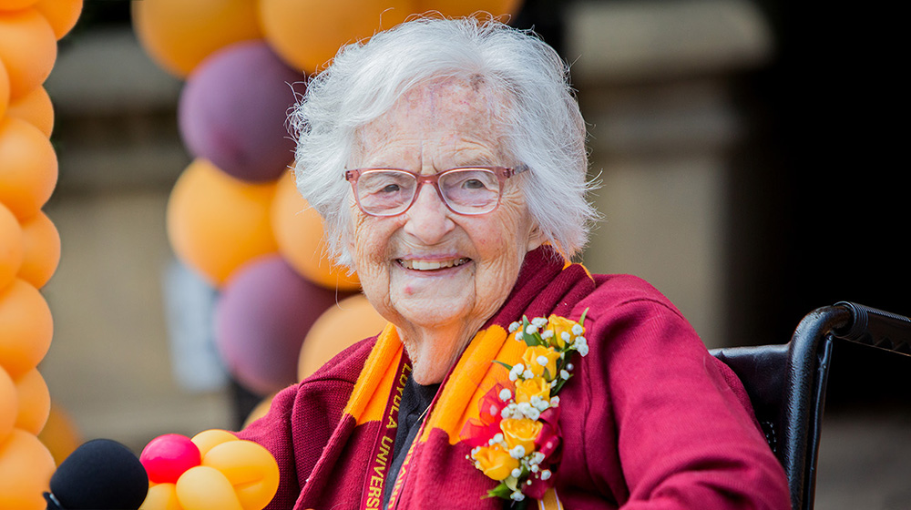 Sister Jean wears a maroon blazer with a five yellow rose corsage on the label and a striped maroon and gold scarf draped over the back of her neck. Sister Jean wears a maroon blazer with a five yellow rose corsage on the label and a striped maroon and gold scarf draped over the back of her neck.