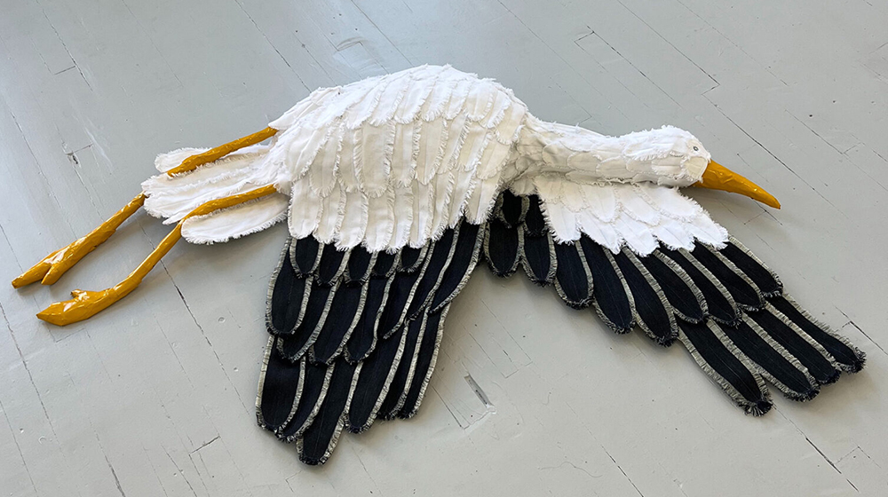 A dimensional, glossy, sculpture of a white stork with black tipped wings in a flying position is displayed on a gray wall. A dimensional, glossy, sculpture of a white stork with black tipped wings in a flying position is displayed on a gray wall.