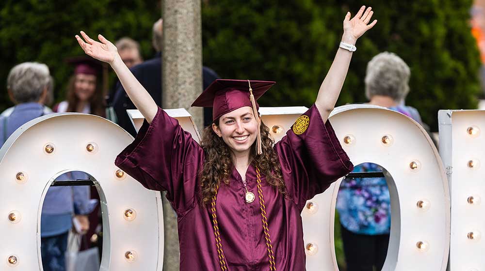 A student in maroon graduation robes lifting her arms in joy A student in maroon graduation robes lifting her arms in joy