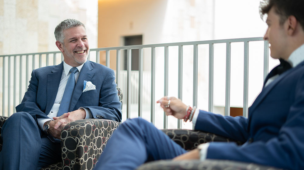 Eric Holler in a blue suit sitting with a student inside the Schreiber Center. Eric Holler in a blue suit sitting with a student inside the Schreiber Center.